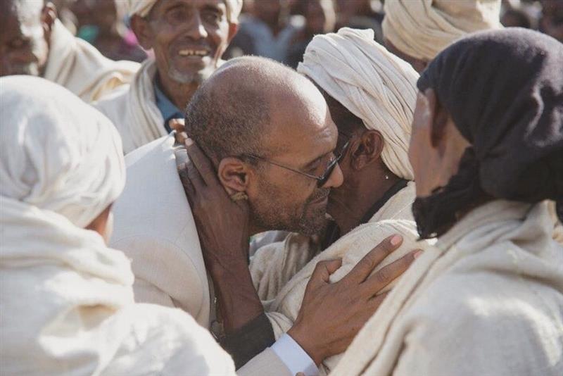 Adugnaw Worku, eyes closed, embraced by elders in white woven gabis in bright highland sunlight on his return home